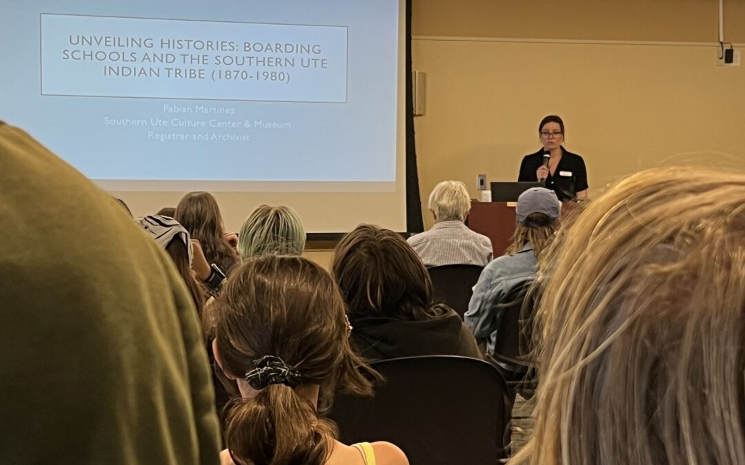 Women standing at podium next to a screen for the "Unveiling Histories: Boarding Schools and the Southern Ute Indian Tribe" presentation.