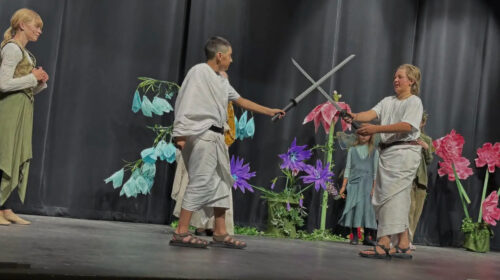 Five PPOS students on stage in costume for a scene from a Midsummers Night Dream. The two characters in center stage are involved in a sword fight.